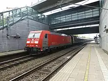 Et moderne rødt og gråt elektrisk lokomotiv trækker et godstog gennem en moderne station af beton og glas. Lokomotivet har førerrum i begge ender, og fremstår lettere kantet men med afrundede ender.