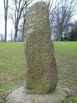 En noget formløs men rektangulær granitsten på en slidt græsplæne. Stenen er indgraveret med runer, som er lidt utydelige grundet den noget ru overflade.