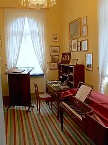 room furnished in early nineteenth century style, with striped runner on floor; walls painted peach and wall on right hung with small pictures; in the right foreground a square piano, in left rear before a window a reading stand, in right rear a desk, all in dark wood