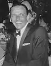 A middle-aged man in a tuxedo sits and smiles into the distance.
