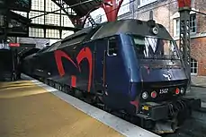 Et blåt, kantet lokomotiv, med førerrum i begge ender og et stort rødt vingehjul med én vinge på siden, er på vej ind til perronen. Perronen befinder sig inde i en stor buehal af rødt og gråt træ stående på grønne stålgittermaster. Buehallens ende over sporet er lukket med mange små matterede glasruder.