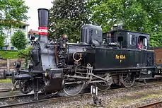 Lille sort damplokomotiv, med rødt og hvidt skorstensbånd, holder foran et par træer. Der er fyret op i lokomotivet og der kommer damp ud ved cylindrene.