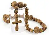Et halsbånd af let irregulære trækugler med et lille, udskåret trækors i midten, vist oven på en gennemsigtig plasticcylinder mod en hvid baggrund