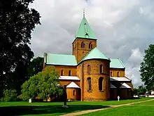A cruciform, copper-roofed, brick church in Romanesque style with rounded window arches and domed apse. The large tower has pointed Gothic-styled windows.
