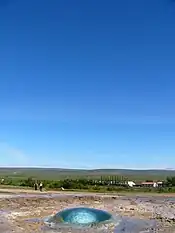 Circle of water in the ground with houses and a green skyline in the distant background.