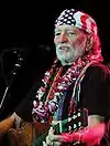 An elderly man holds a guitar while in front of a microphone. He has white facial hair and wears a United States flag bandana.
